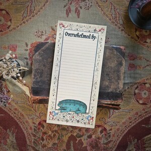 May include: A lined notepad with the words "Overwhelmed by:" and a blue frog illustration. The notepad is decorated with floral and scroll designs. It is placed on a stack of old books, with a decorative background.