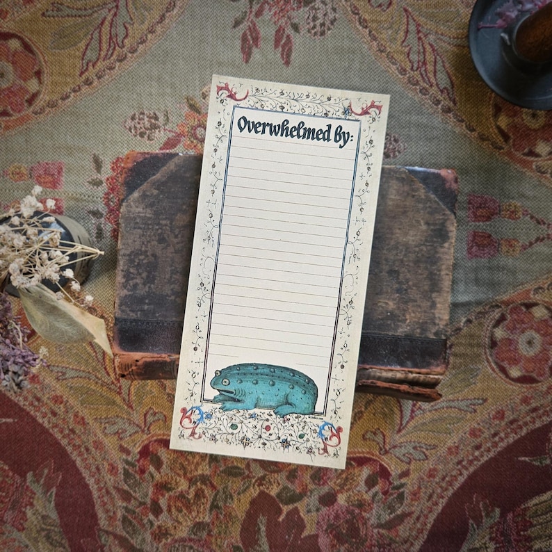 May include: A lined notepad with the words "Overwhelmed by:" and a blue frog illustration. The notepad is decorated with floral and scroll designs. It is placed on a stack of old books, with a decorative background.