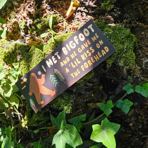 Puede incluir: Una pegatina rectangular con fondo oscuro que presenta una ilustraci&oacute;n de un Bigfoot marr&oacute;n, pinos verdes y el texto: I MET BIGFOOT AND HE GAVE ME A 'LIL KISS ON THE FOREHEAD. El dise&ntilde;o incluye peque&ntilde;as nubes amarillas y una estrella blanca.