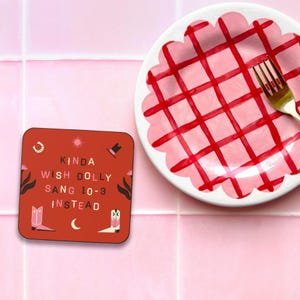 Pu&ograve; includere: Un sottobicchiere quadrato con sfondo rosso e testo bianco che recita "Kinda wish Dolly sang 10-3 instead". Il sottobicchiere ha un ferro di cavallo, un sole, un cappello da cowboy, stivali e una luna crescente negli angoli.