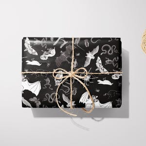 May include: A wrapped gift with black wrapping paper featuring a white and gray pattern of bats, snakes, and other creatures. The gift is tied with twine and a bow. A pair of black scissors and a spool of twine are also in the image.