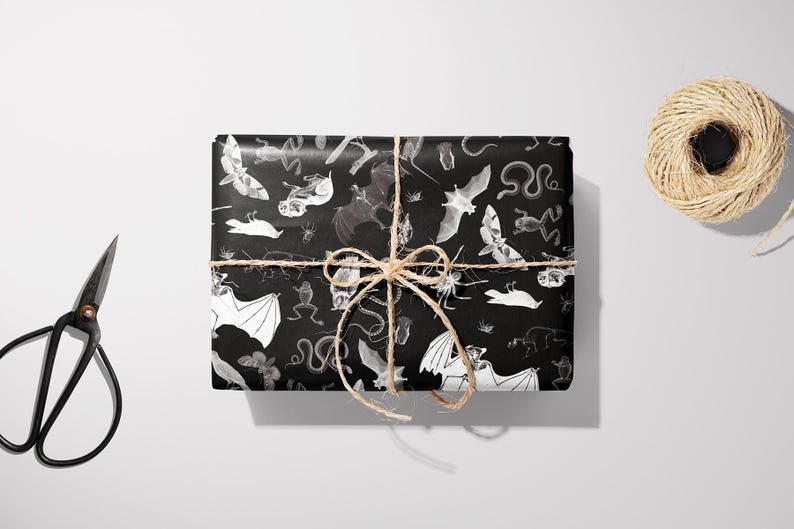 May include: A wrapped gift with black wrapping paper featuring a white and gray pattern of bats, snakes, and other creatures. The gift is tied with twine and a bow. A pair of black scissors and a spool of twine are also in the image.