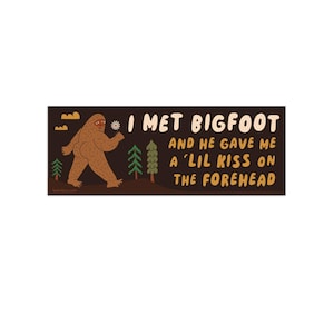 Puede incluir: Una pegatina rectangular con fondo marr&oacute;n oscuro que muestra una ilustraci&oacute;n de un Bigfoot marr&oacute;n, pinos verdes y nubes amarillas. El texto dice: I MET BIGFOOT AND HE GAVE ME A 'LIL KISS ON THE FOREHEAD en letras color crema y amarillo.