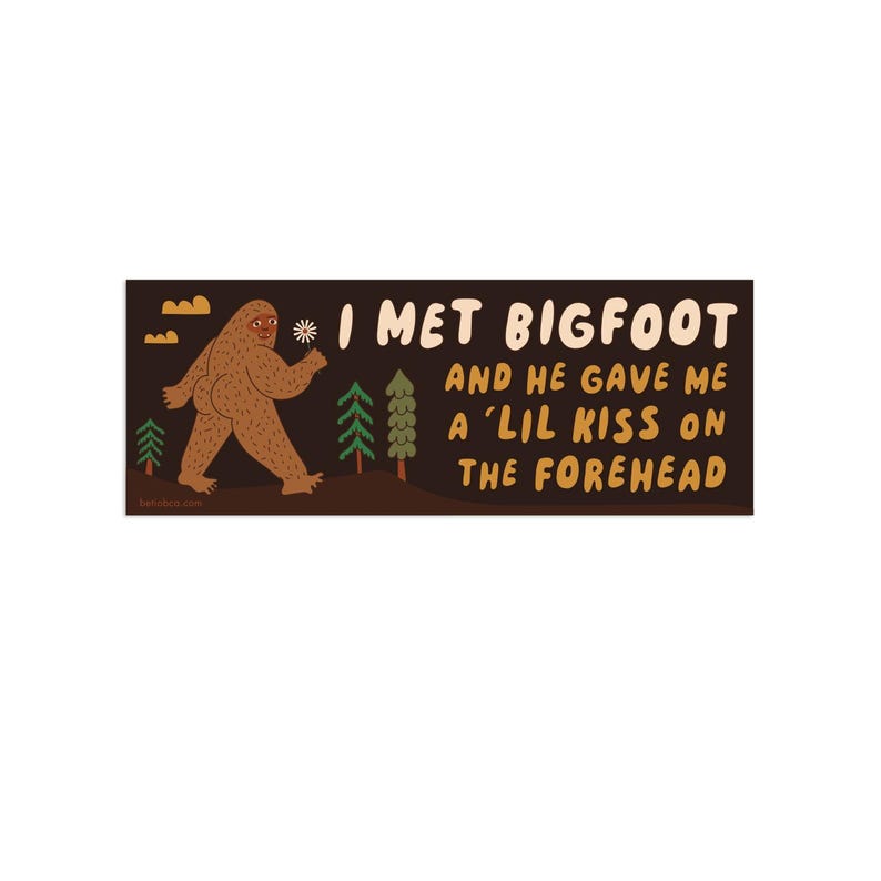 Puede incluir: Una pegatina rectangular con fondo marr&oacute;n oscuro que muestra una ilustraci&oacute;n de un Bigfoot marr&oacute;n, pinos verdes y nubes amarillas. El texto dice: I MET BIGFOOT AND HE GAVE ME A 'LIL KISS ON THE FOREHEAD en letras color crema y amarillo.