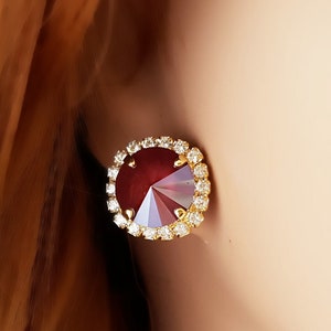 May include: A pair of gold stud earrings with a red rhinestone center surrounded by a halo of smaller clear rhinestones.