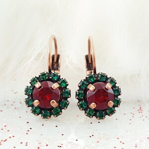 May include: A pair of leverback earrings featuring a central round red stone surrounded by a halo of small green crystals. The metal setting has a warm copper tone, and the design includes four prongs securing the large red center stone.