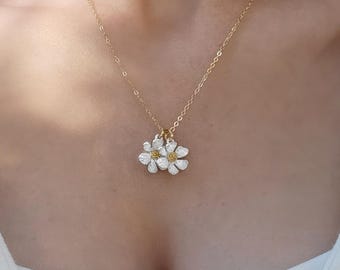 May include: A gold-toned chain necklace featuring two small, white daisy-shaped charms with yellow centers. The necklace rests against a white ribbed top. The delicate metal chain connects to the floral pendants, which hang symmetrically at the center.