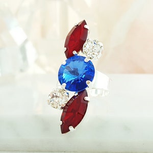 May include: A silver ring with a blue, red, and clear crystal design. The ring features a large blue crystal in the center, surrounded by two red crystals and two clear crystals.