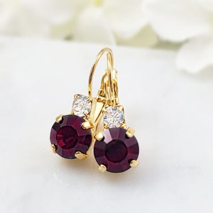 May include: A pair of gold earrings with a deep burgundy round gemstone and a smaller clear rhinestone accent.