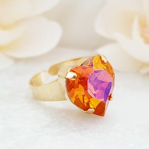 May include: A gold-toned adjustable ring featuring a large, heart-shaped crystal. The crystal displays a vibrant color gradient of orange, yellow, and purple hues. The ring's band is simple and elegant, with a polished finish.