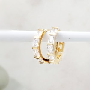 May include: A pair of gold hoop earrings with a row of clear square-shaped cubic zirconia stones around each hoop.