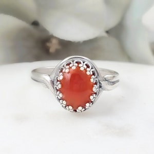May include: A silver ring with a red coral stone set in a delicate, ornate bezel. The ring has a simple band with a slight curve.