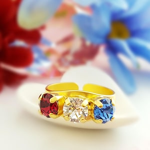 May include: A gold-toned adjustable ring featuring three round gemstones: a red ruby, a clear diamond, and a blue sapphire. The ring is set on a white heart-shaped surface, with a blurred floral background in red and blue.