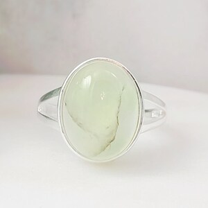 May include: A silver ring with a light green oval-shaped gemstone. The ring is set in a simple, elegant design.