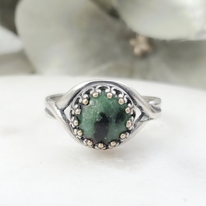May include: A silver ring with a green gemstone set in a gold bezel. The ring has a delicate, twisted band.