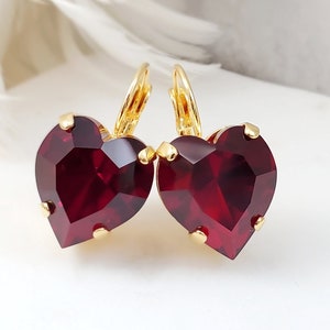 May include: A pair of heart-shaped earrings with faceted, deep red crystals. The earrings have gold-colored lever-back closures and gold-colored prongs holding the crystals. The earrings are displayed on a white surface with a soft, blurred background.