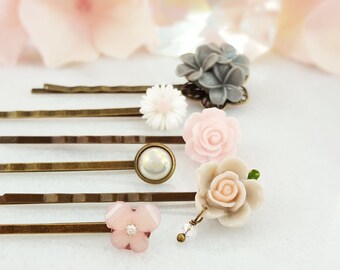 Puede incluir: Conjunto de seis pinzas para el pelo doradas con diversos adornos florales, incluyendo rosas rosas, una margarita blanca, una flor gris y una flor rosa con un centro de pedrería.