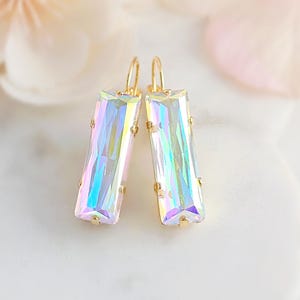 May include: A pair of rectangular earrings with a gold-colored frame and iridescent, rectangular crystals. The crystals reflect a rainbow of colors. The earrings are displayed on a white surface with soft, blurred floral elements in the background.