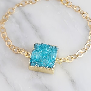 May include: Gold-toned chain bracelet featuring a square, turquoise-colored druzy stone. The stone is set in a gold-colored bezel. The bracelet has an adjustable length and a lobster clasp closure. Jewelry.