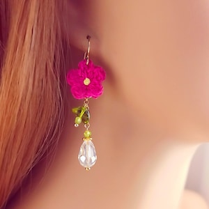 May include: A handmade earring featuring a vibrant fuchsia crocheted flower with a gold center. Below the flower, green leaves and beads dangle, leading to a clear, teardrop-shaped crystal. The earring has a gold-colored hook.