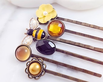 May include: A set of six gold hair clips with various decorative charms, including a bee, a flower, and round gems in black, yellow, and amber.