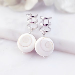May include: A pair of silver-toned ear cuffs with dangling white, spiral-patterned pendants. The earrings have a modern design, with a polished finish. The white pendants have a subtle, natural swirl pattern. The earrings are displayed on a white surface.