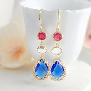 May include: A pair of gold-tone drop earrings featuring a vertical arrangement of three faceted stones. The top stone is round and pink, the middle is oval and clear, and the bottom is a large, pear-shaped blue stone with a decorative gold border.