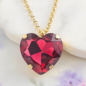 May include: A gold-toned necklace featuring a large, heart-shaped pendant. The pendant is a faceted, deep red gemstone, set in a gold bezel. The chain is delicate and also gold-colored, creating a romantic and elegant piece of jewelry.
