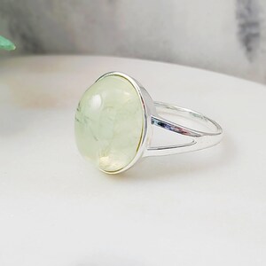May include: A silver ring with a light green oval gemstone. The ring has a simple design with a split band.