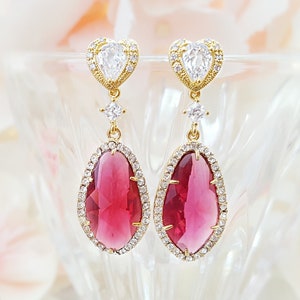 May include: A pair of gold-toned drop earrings with a heart-shaped top, a small square gem, and a large, teardrop-shaped, ruby-red gem surrounded by small clear gems. The earrings are displayed in a clear glass bowl with a soft, floral background.