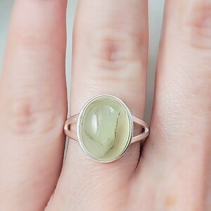 May include: A silver ring with a light green oval gemstone. The ring is on a finger.