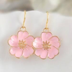 Puede incluir: Un par de pendientes de flores dorados con pétalos de esmalte rosa y un centro dorado. Los pendientes tienen forma de flor con un diseño delicado. Se muestran sobre una superficie blanca.