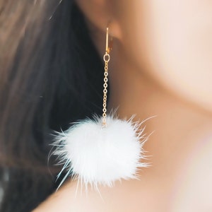 May include: A dangling earring featuring a gold-toned metal chain attached to a small lever-back hook. At the base of the chain hangs a single, round, fluffy white pom-pom made of soft, fine fibers that create a textured, spherical appearance.
