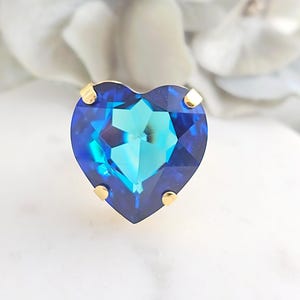 May include: A heart-shaped ring featuring a faceted deep blue and teal gemstone set in a gold-toned metal prong setting. The stone displays a vibrant color gradient with a brilliant cut that reflects light across its polished surface.