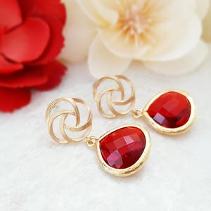 May include: Gold-toned earrings featuring a woven knot design and teardrop-shaped, faceted red gemstones. The earrings have a gold frame around the red stones. The background includes red and white flowers.