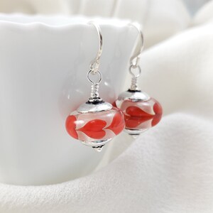 May include: A pair of silver dangle earrings with red heart-shaped glass beads. The beads are translucent and have a white background.