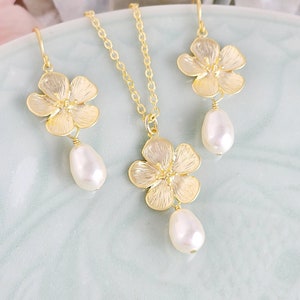 May include: A gold necklace and earrings set featuring delicate flower charms with pearl drops. The flowers have a textured, almost hammered, finish.
