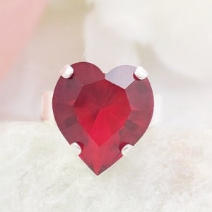 May include: A silver-toned ring featuring a large, faceted, heart-shaped deep red gemstone. The stone is secured by four small metal prongs at the top, bottom, and sides, creating a prominent centerpiece on the band of the ring.