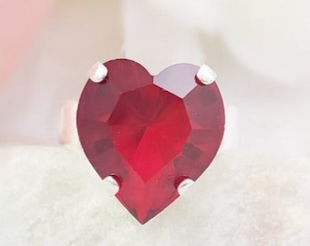 May include: A silver-toned ring featuring a large, faceted, heart-shaped deep red gemstone. The stone is secured by four small metal prongs at the top, bottom, and sides, creating a prominent centerpiece on the band of the ring.