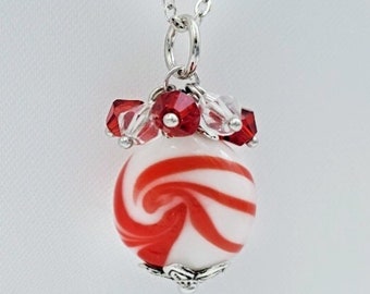 May include: A silver chain necklace with a red and white candy cane swirl glass bead pendant. The pendant is accented with clear and red crystal beads.