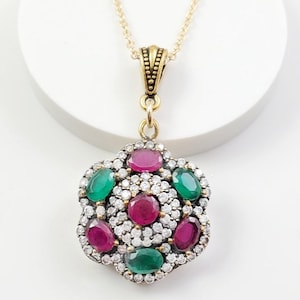 May include: A gold-toned necklace featuring a flower-shaped pendant with oval-cut red and green stones surrounded by small white crystals. The pendant is suspended from a delicate gold chain. The text Style 5 is visible in the corner.
