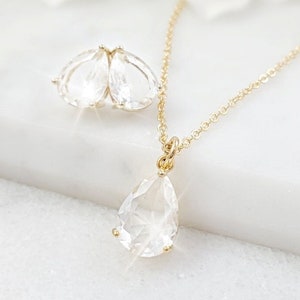 Puede incluir: Un collar de oro con un colgante de cristal en forma de l&aacute;grima y un par de pendientes a juego con l&aacute;grimas de cristal. Las joyas est&aacute;n sobre una superficie blanca.