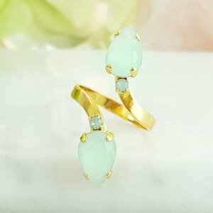 May include: A gold ring with two teardrop-shaped green stones and smaller clear stones. The ring is designed to wrap around the finger.
