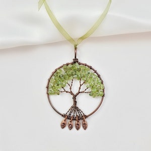 May include: A green peridot stone tree of life pendant with copper wire branches and roots. The pendant is hanging from a green ribbon and has the year 2022 inscribed on the leaves.
