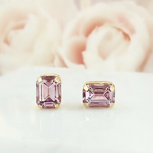 May include: Two gold-toned, emerald-cut stud earrings with a light purple gemstone. The earrings are set on a white surface, with soft pink and purple floral elements in the background. The earrings are a classic jewelry item.