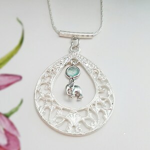 May include: A silver necklace with a teardrop-shaped pendant featuring an intricate filigree design. The pendant has a small light blue gemstone and a silver elephant charm hanging from it.