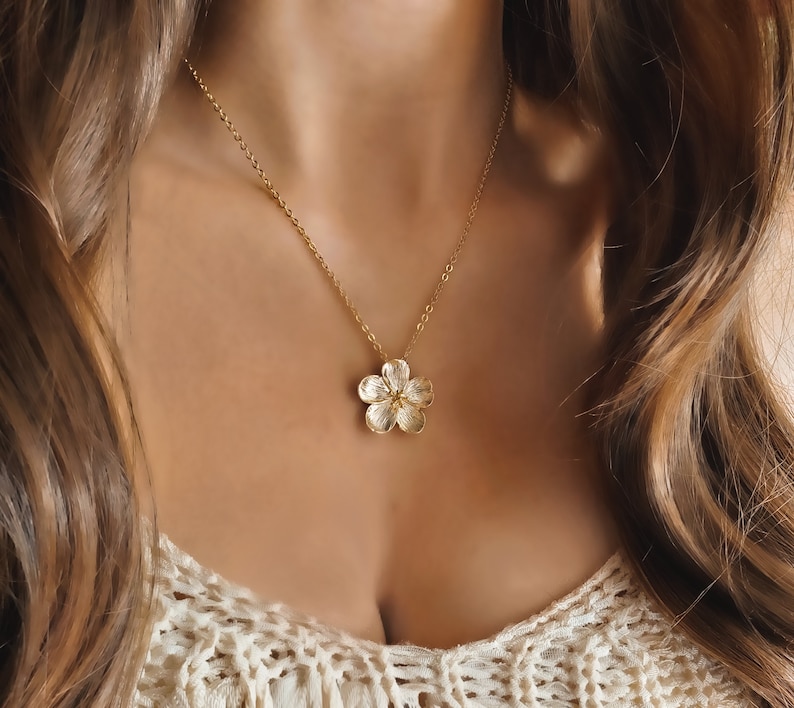 May include: A gold chain necklace with a delicate flower pendant. The flower is textured and has a slightly raised centre.