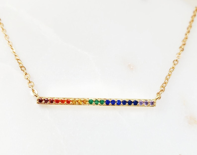 May include: A gold necklace with a thin bar pendant featuring a rainbow of colorful gemstones.