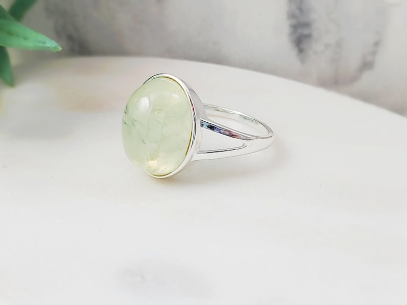 May include: A silver ring with a light green oval gemstone. The ring has a simple design with a split band.