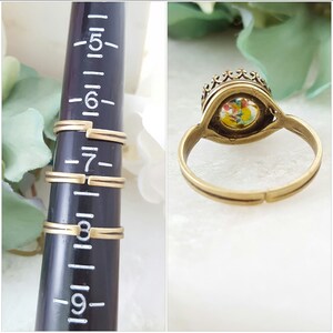 May include: A gold-toned ring with a crown-shaped setting. The setting contains a colorful, speckled stone. The ring is shown on a sizing tool, indicating sizes 5 through 9.